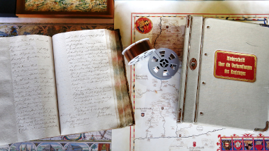 Bild: Aufgeschlagenes Buch mit Handschrift, geschlossenes Buch mit rot goldener Beschriftung, Mikofilmrollen und Kartenmaterial