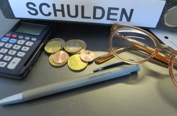 Ordner mit der Aufschrift "Schulden", Taschenrechner, Geldmünzen, Brille und Kugelschreiber