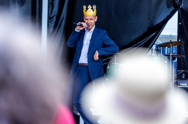 Ein Mann im Anzug steht auf einer Bühne mit einem Mikro in der Hand und einer Krone Auf dem Kopf