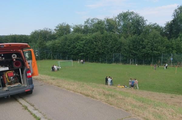 ©Vermessung und Flurneuordnung/Landratsamt Zollernalbkreis Links im Bild steht der orangene Vermessungsbus, durch die offene Kofferraumtüre sind Vermessungsgerätschaften zu sehen. Mittig und rechts im Bild sind viele Schüler an verschiedenen Stationen auf einer Wiese zu sehen.