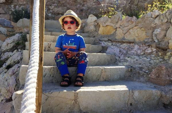 Lange, weite Kleidung, Kopfbedeckung: Kinder benötigen bei Hitze und Sonne besonderen Schutz. ©Landratsamt Zollernalbkreis Ein Bub sitzt auf einer Treppe, er trägt einen Strohhut, Sonnenbrille