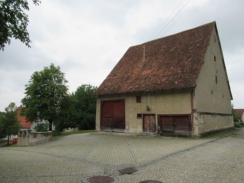 Eine alte Scheune, davor ein gepflasterter Hof und daneben Bäume.