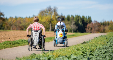 Zwei Personen in Rollstühlen auf einem Feldweg